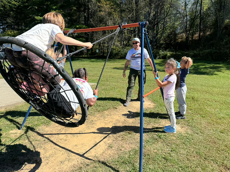 Children laugh and play on a large net swing while a Project:Camp volunteer watches nearby on a sunny day in Asheville.