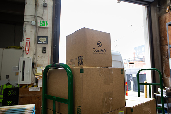 A Good360-branded cardboard box sits on a hand truck at a loading dock, ready for delivery to WellPower in Denver, Colorado.